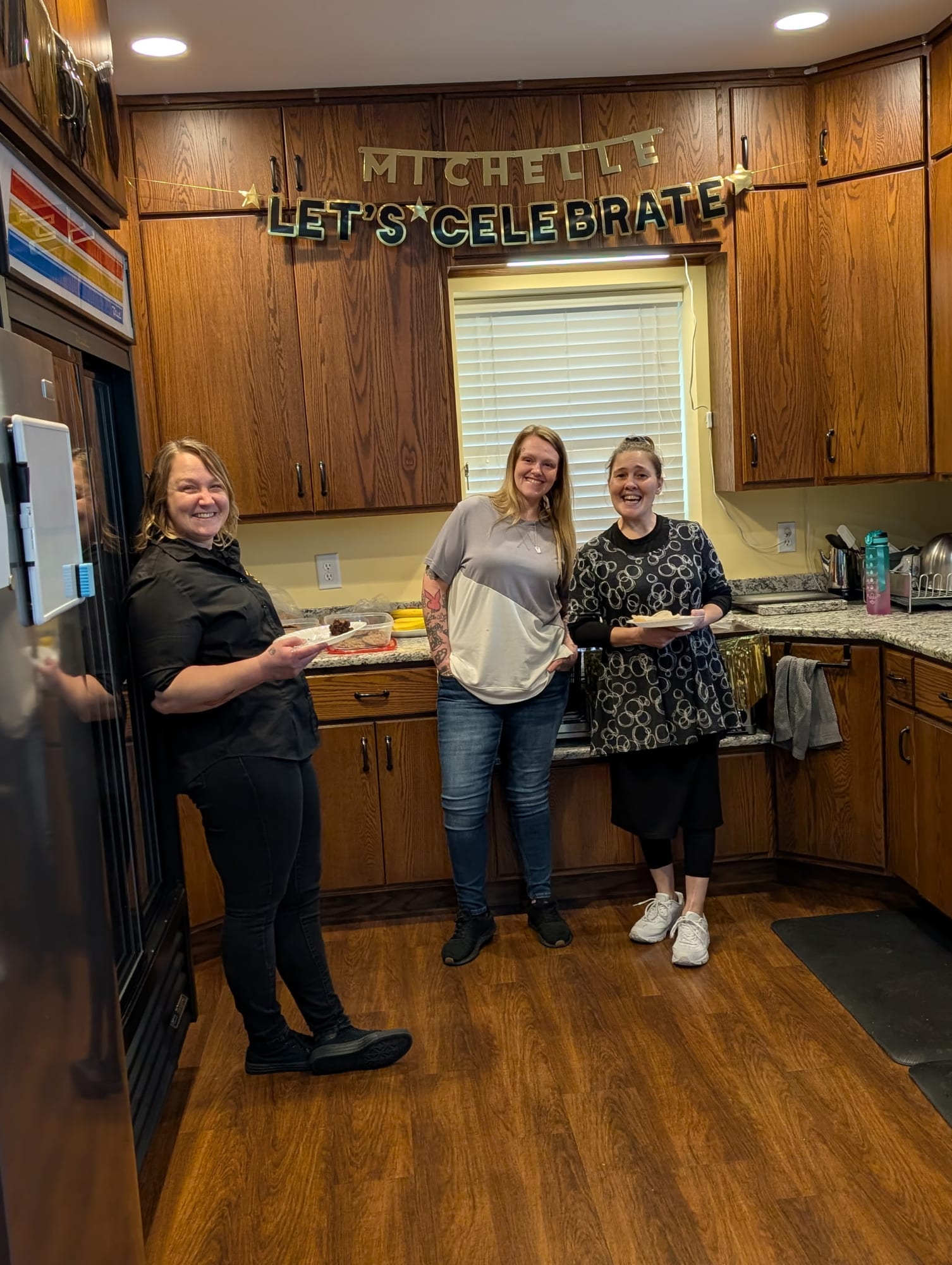 Three women with plates of food in the kitchen, under a Let's Celebrate banner.
