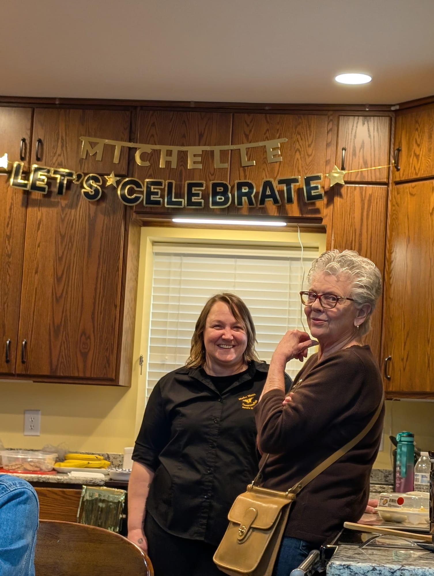 Two women smiling in the kitchen under a celebration banner.