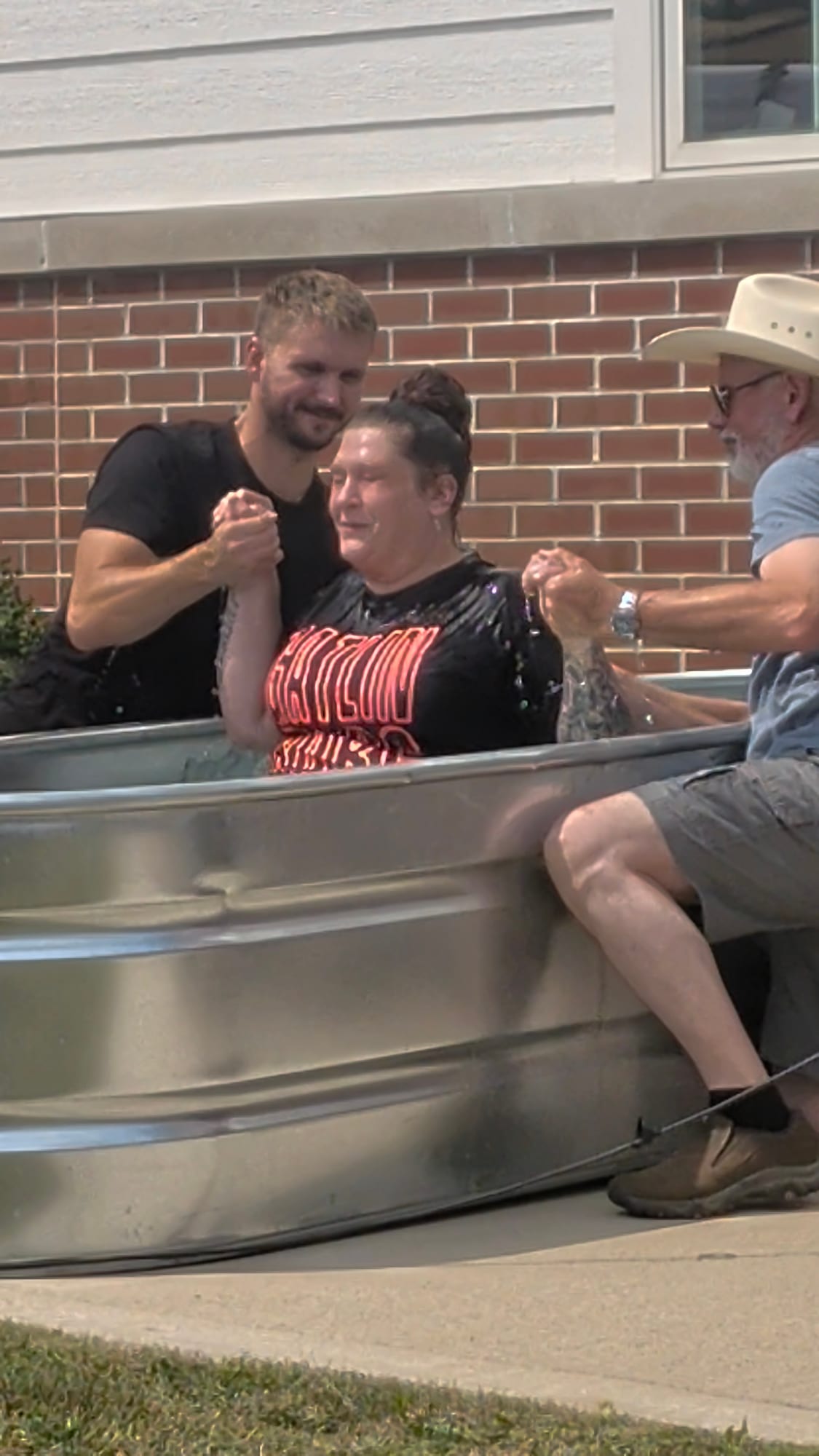 A woman being baptized in a stock tank outside a church, held by two men.